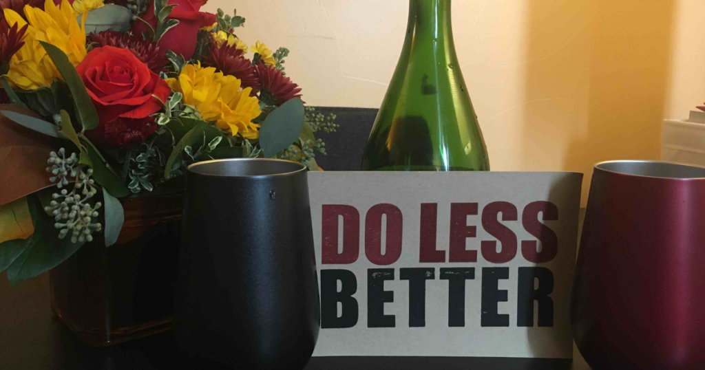 Two wine glasses with a sign that says "do less better" in between. There is a flower arrangement on the left and a bottle of wine behind the sign. This represents our new outlook to help you, something we've thought about for a while it turns out.