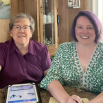 Two woman smiling, sitting at a table, with an iPad in front of them.